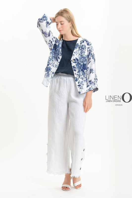 Against a white background, a person wears the French Linen Jacket TP1523P-11 Bianco by LinenO with a dark blue top, white wide-legged pants, and white sandals. Made from eco-friendly French linen; they stand relaxed with one hand on their forehead. The "LINEN ORIGINS" logo is visible on the right.
