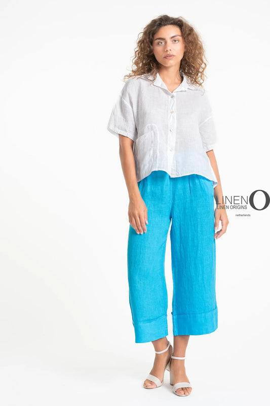 A woman wearing a French Linen Shirt TP1575-11 Bianco from LinenO and bright blue wide-legged pants stands against a white background. Her look is completed with eco-friendly white strappy sandals, and her naturally wavy hair complements her relaxed style.