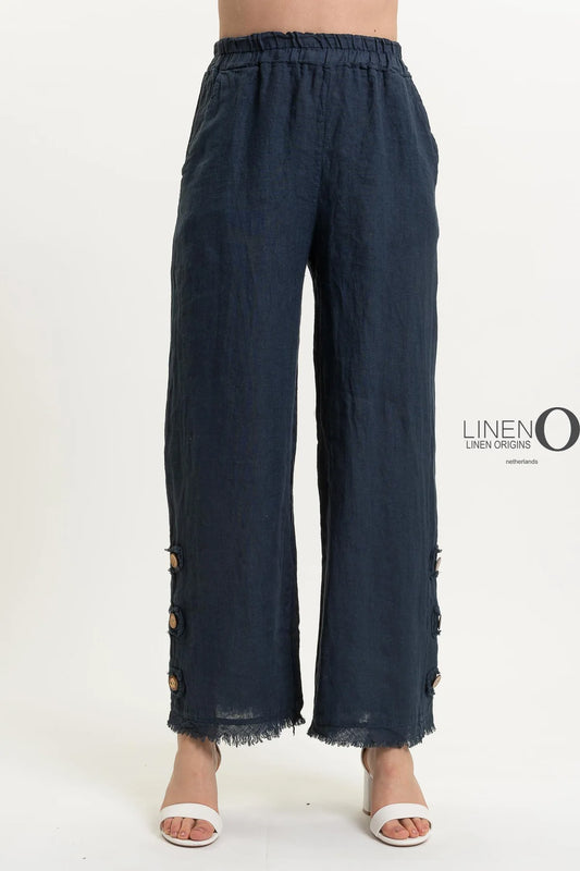 A person wears the stylish French Linen Pant PT839-75 Notte by LinenO, featuring wide-leg dark blue European linen with frayed hem and wooden buttons near the bottom. Paired with white open-toed heels on a crisp white background, it highlights eco-friendly fashion choices.