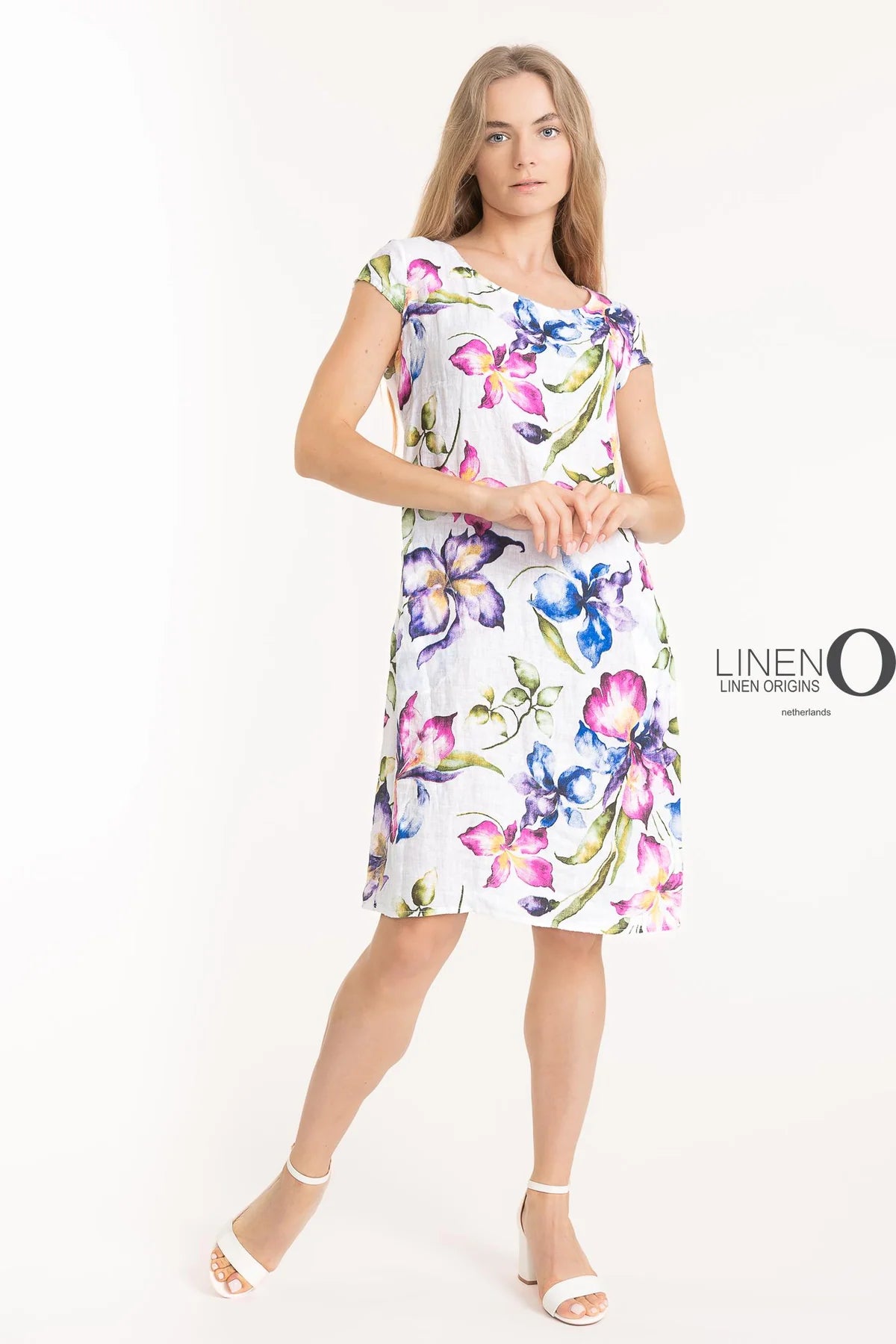 A person in a knee-length French Linen Dress TP1511-11 Bianco by LinenO, with vibrant purple, pink, and blue floral patterns, stands against a light background. They're wearing white sandals. "Linen Origins" is featured on the right side, showcasing the charm of European linen.