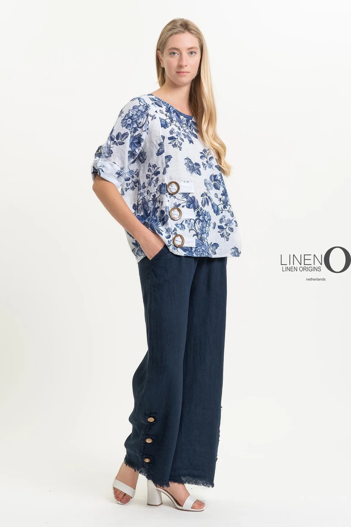 A woman wearing the "French Linen Tunic TP1518P-11 Bianco" by LinenO stands against a light background. Her floral-patterned blouse and dark wide-leg pants, both crafted from 100% European linen, exude eco-friendly elegance. The word "LINEN" is prominently displayed to highlight sustainable French linen fashion.