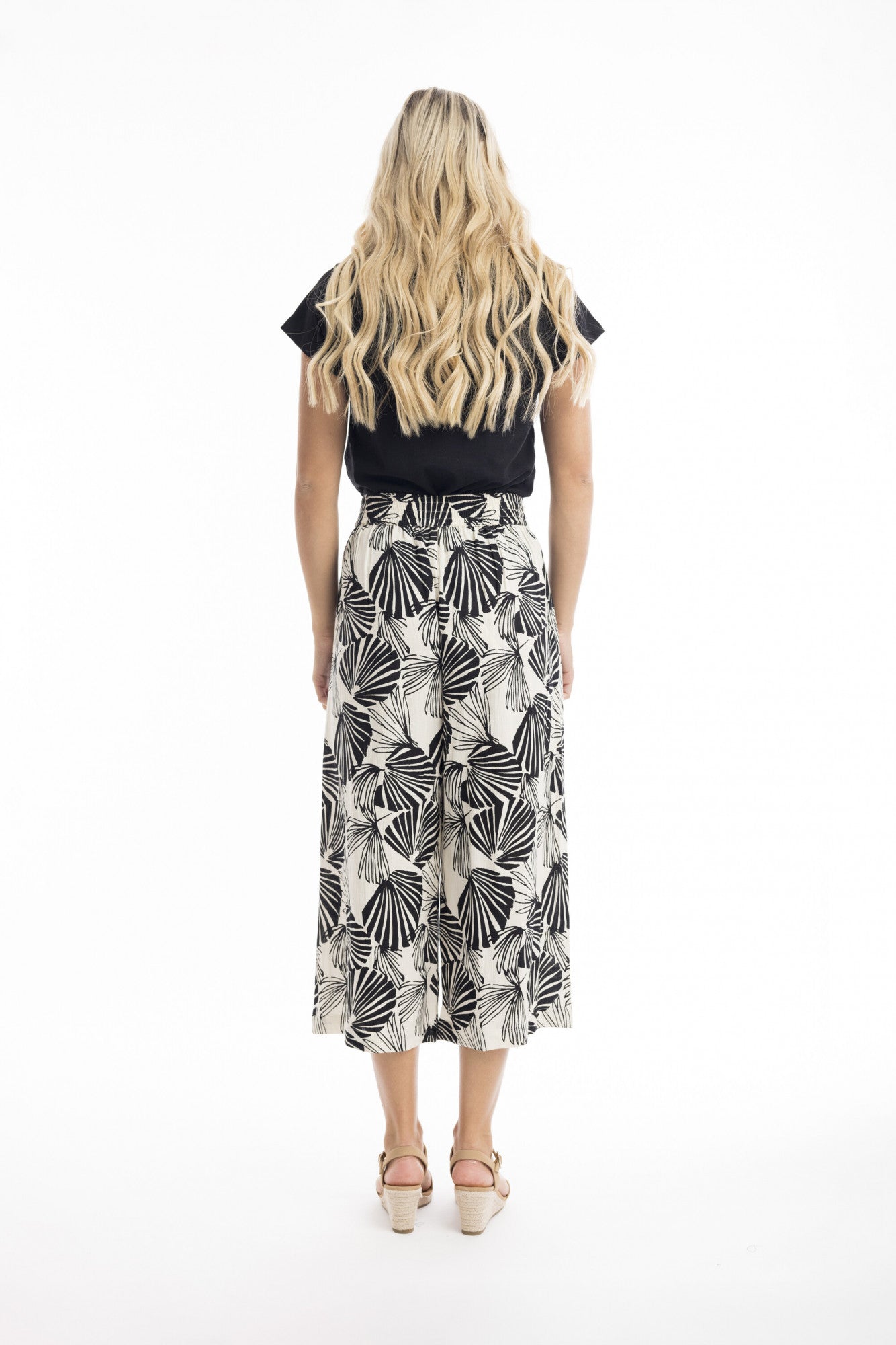 A woman with long, wavy blonde hair stands facing away, wearing ORIENTIQUE's Papagayo Pants Style: 76048—wide-leg linen blend pants with a black and white geometric leaf pattern—plus a black top and light wedge sandals against a white background.
