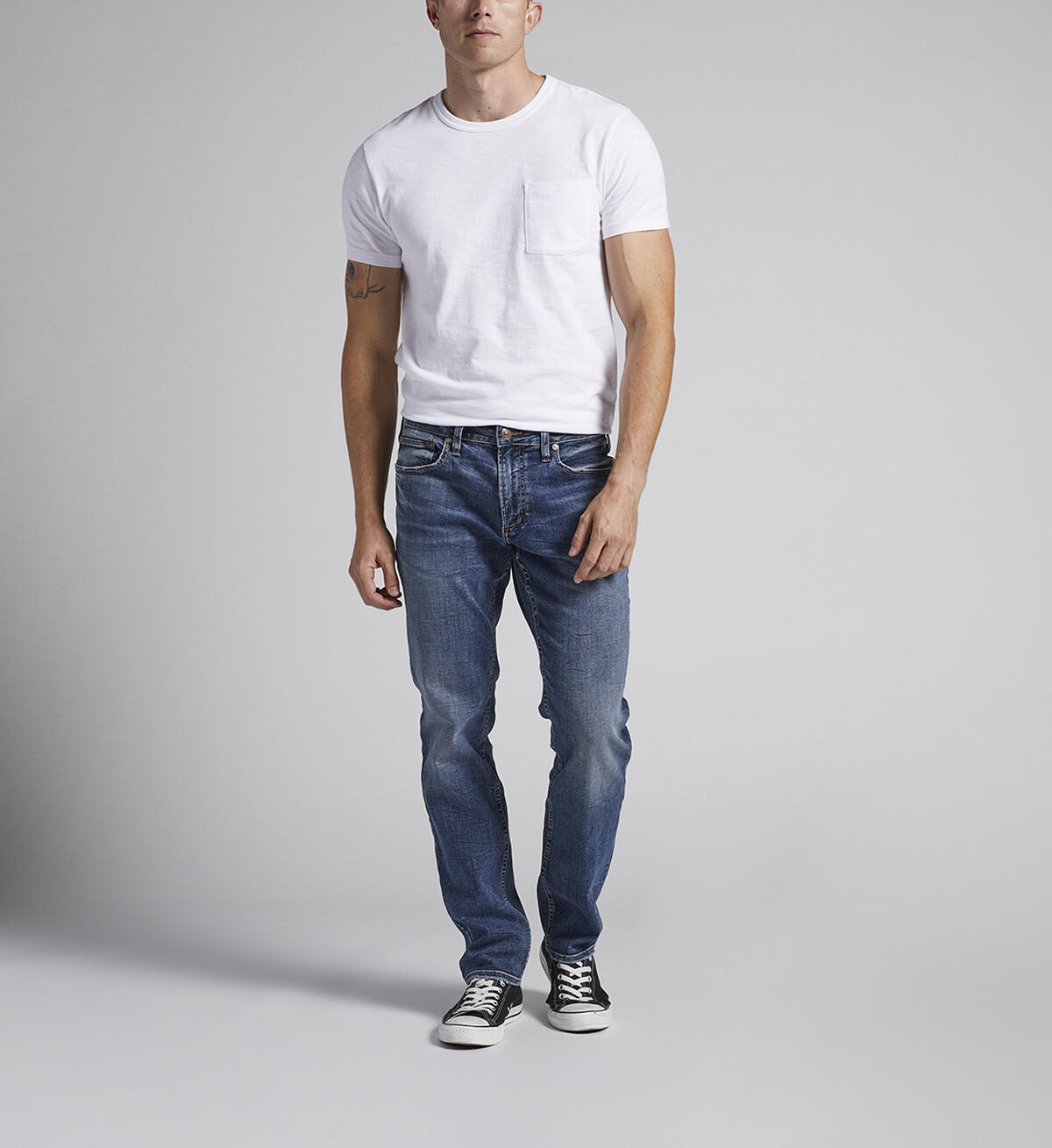 A man is standing against a plain grey background, wearing a white T-shirt, Konrad Slim Fit Slim Leg Jeans from Silver Jeans Co., and black sneakers. His hands are relaxed by his sides, and he is facing slightly to the right. Only the lower part of his face is visible.