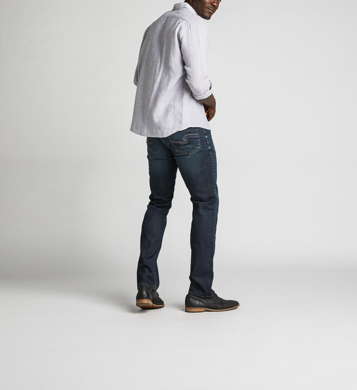 A man stands against a plain background, wearing a light gray button-up shirt with rolled-up sleeves, Konrad Slim Fit Slim Leg Jeans from Silver Jeans Co. in dark indigo rinse, and dark brown shoes. He looks towards the camera with a relaxed posture, with one hand slightly in front.