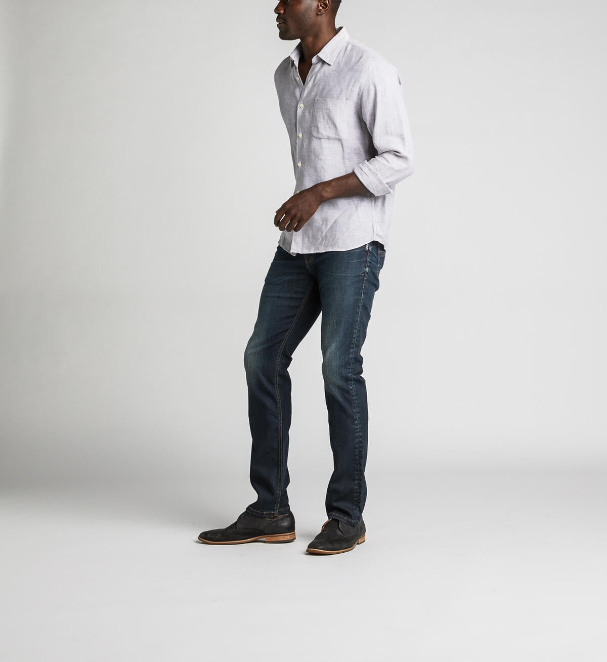 A man stands against a plain background, wearing a light gray button-up shirt with rolled-up sleeves, Konrad Slim Fit Slim Leg Jeans from Silver Jeans Co. in dark indigo rinse, and dark brown shoes. He looks towards the camera with a relaxed posture, with one hand slightly in front.