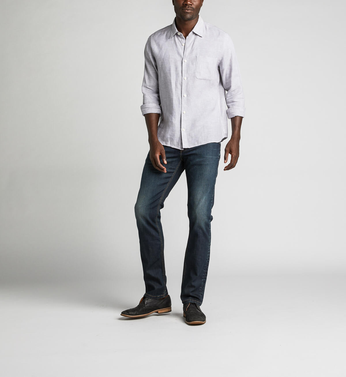 A man stands against a plain background, wearing a light gray button-up shirt with rolled-up sleeves, Konrad Slim Fit Slim Leg Jeans from Silver Jeans Co. in dark indigo rinse, and dark brown shoes. He looks towards the camera with a relaxed posture, with one hand slightly in front.