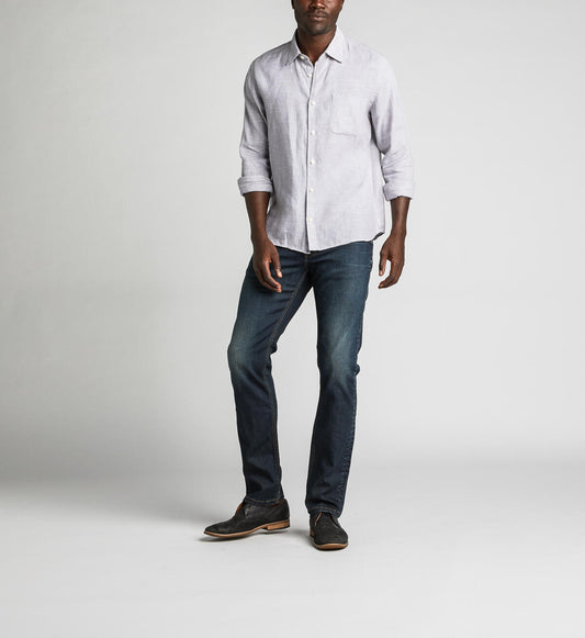 A man stands against a plain background, wearing a light gray button-up shirt with rolled-up sleeves, Konrad Slim Fit Slim Leg Jeans from Silver Jeans Co. in dark indigo rinse, and dark brown shoes. He looks towards the camera with a relaxed posture, with one hand slightly in front.