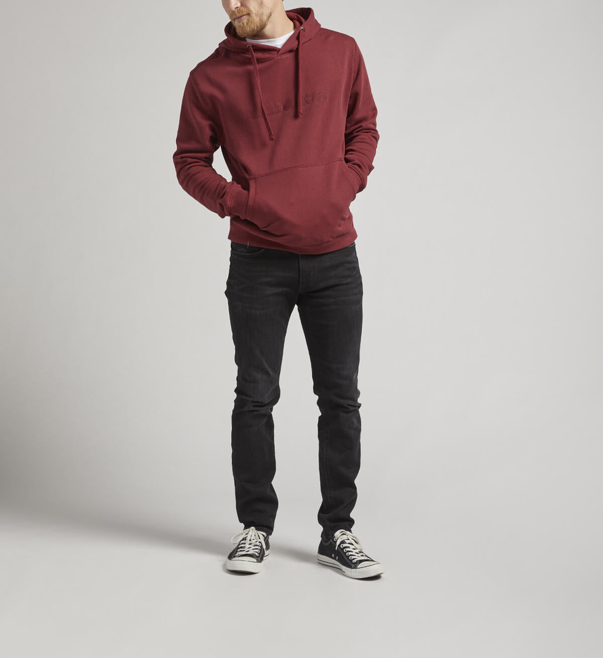 A person with short hair stands facing away from the camera, wearing a Silver Jeans Co. Logo Hoodie in burgundy featuring an adjustable drawstring. They pair it with black jeans and black shoes with white soles. The background is a plain, light gray color.