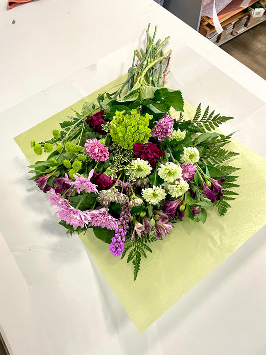 The *Emerald and Violet Delight* by *Posies Flowers & Fashion* features a colorful bouquet of various flowers, including pink, purple, and white blooms, intertwined with lush green foliage. The vibrant arrangement is beautifully spread out on light green wrapping paper laid on a white surface, with some stems elegantly extending beyond the wrapping. Flower substitutions may be made based on availability.