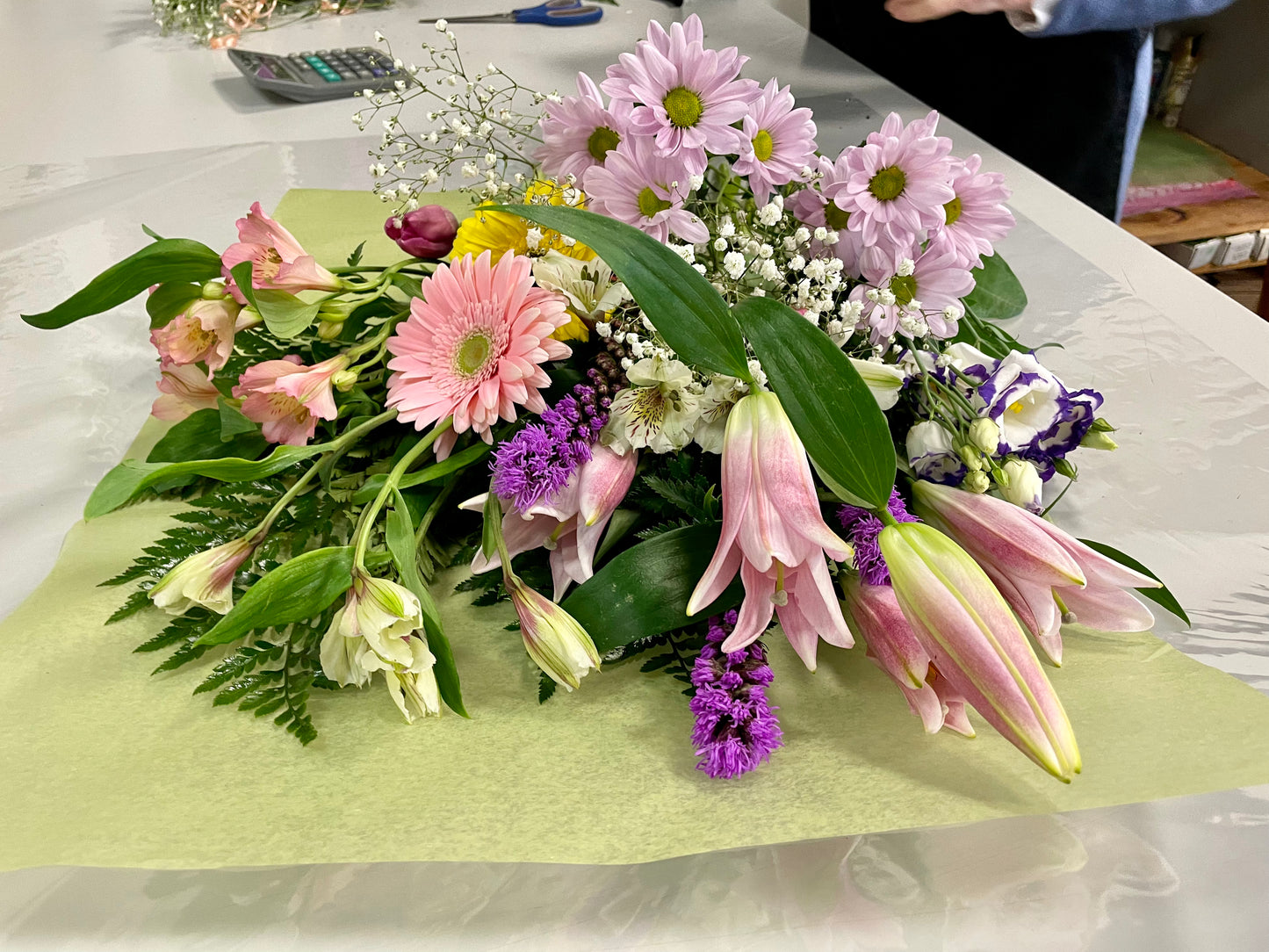 Introducing "Mother’s Delight" by Posies Flowers & Fashion: a vibrant bouquet featuring an exquisite mix of pink lilies, white lilies, pink gerberas, red carnations, yellow flowers, purple flowers, and airy sprigs of baby's breath. The blossoms are beautifully arranged on a light green and white surface and surrounded by lush green leaves. Please note that substitutions may occur based on availability.
