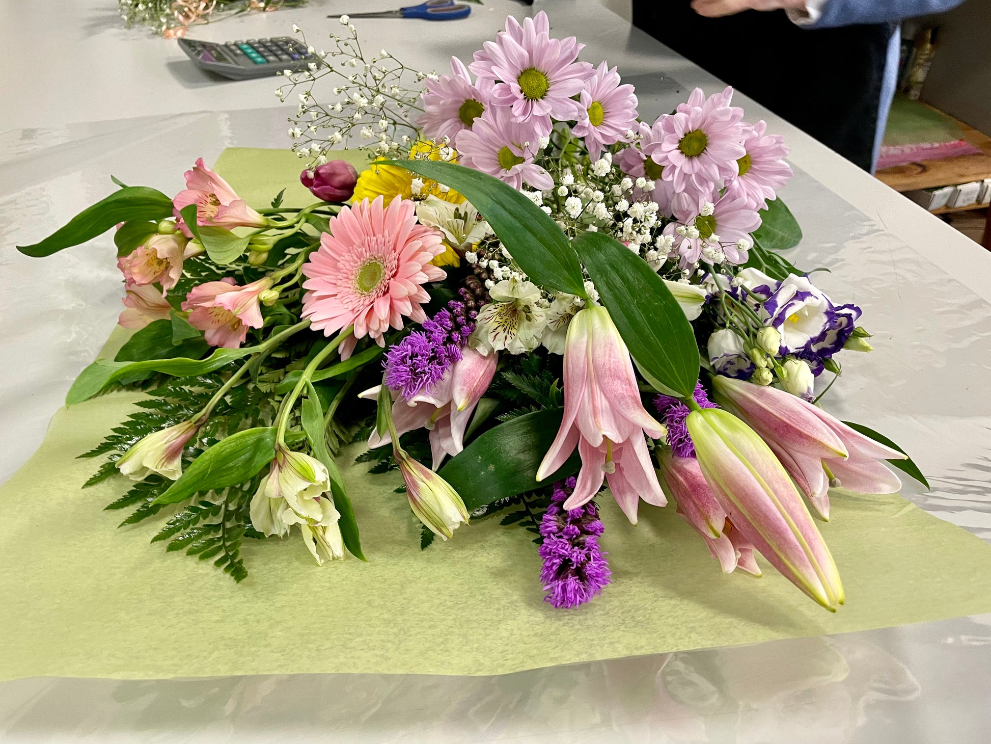 Introducing "Mother’s Delight" by Posies Flowers & Fashion: a vibrant bouquet featuring an exquisite mix of pink lilies, white lilies, pink gerberas, red carnations, yellow flowers, purple flowers, and airy sprigs of baby's breath. The blossoms are beautifully arranged on a light green and white surface and surrounded by lush green leaves. Please note that substitutions may occur based on availability.