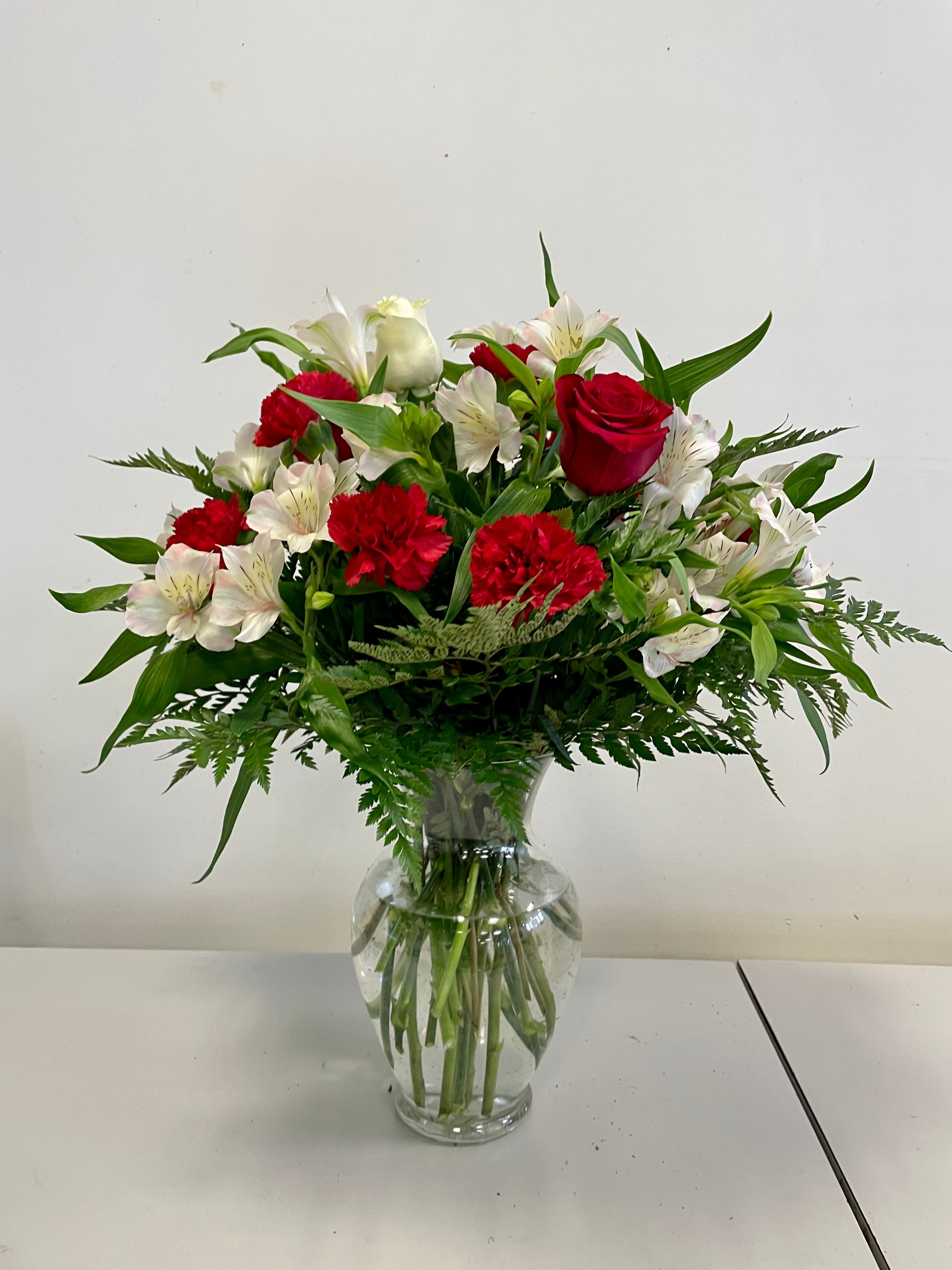The "Love at First Sight" flower arrangement by Posies Flowers & Fashion, featuring a glass vase filled with red roses, red carnations, and white lilies accented with green foliage, sits elegantly on a white surface against a plain white background. The image captures the timeless beauty of these flowers perfectly.