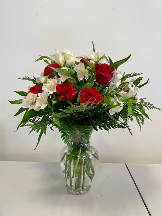 The "Love at First Sight" flower arrangement by Posies Flowers & Fashion, featuring a glass vase filled with red roses, red carnations, and white lilies accented with green foliage, sits elegantly on a white surface against a plain white background. The image captures the timeless beauty of these flowers perfectly.