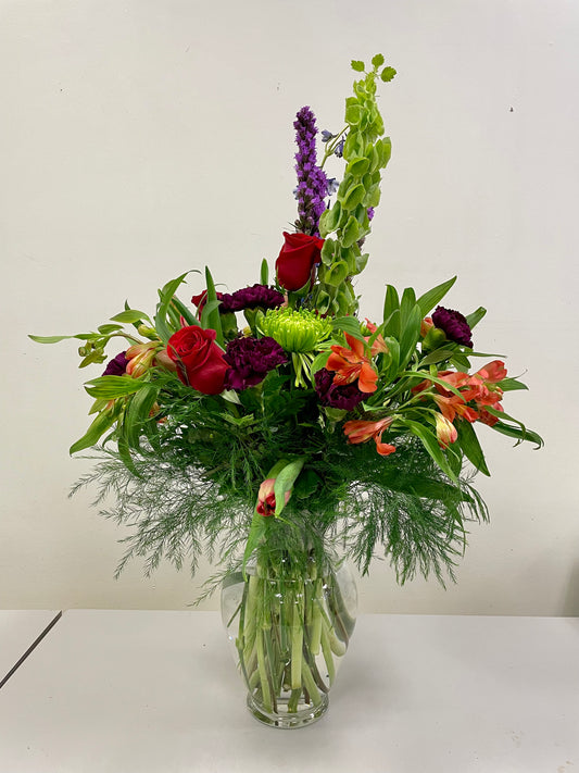Introducing the "Hints of Love" arrangement by Posies Flowers & Fashion: This clear glass vase showcases a beautiful assortment of various flowers, including vibrant red roses, striking orange lilies, delicate green chrysanthemums, and pretty purple statice, all complemented by lush greenery. Displayed against a plain white background for an elegant touch. Please be aware that flower availability may vary, and appropriate substitutes will be provided if needed.