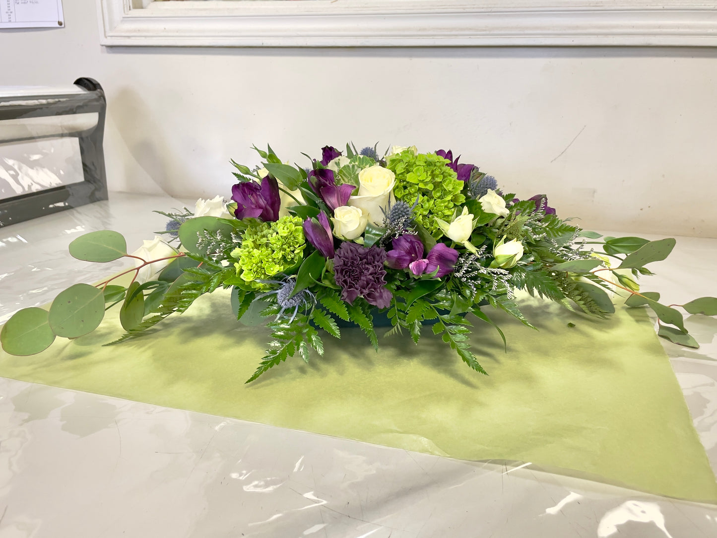 Introducing the "Garden of Royalty Centrepiece" by Posies Flowers & Fashion, showcasing a stunning mix of purple tulips, white roses, and green hydrangeas. Accented with greenery and ferns, this elegant floral arrangement is displayed on a light green placemat atop a white table. This exquisite piece enhances the room's simplicity, against a plain white wall with a partially visible window frame.