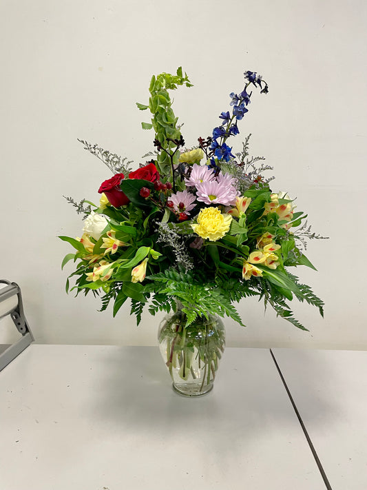The "Love & Wonder" bouquet from Posies Flowers & Fashion is presented in a clear glass vase, showcasing a vibrant assortment of flowers including red roses, yellow carnations, purple chrysanthemums, and blue delphiniums. Accented with green foliage and ferns for an accurate depiction, this stunning arrangement is displayed on a white table against a plain white wall.