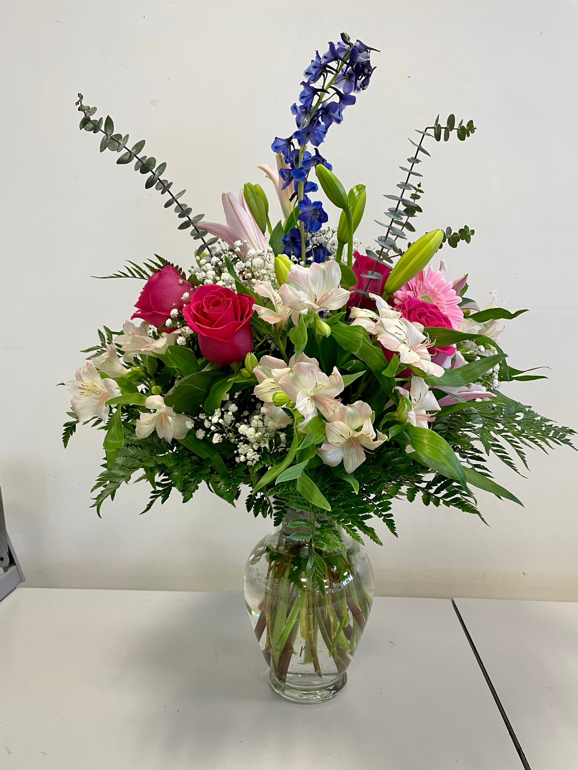 The "Floral Hugs" arrangement by Posies Flowers & Fashion showcases a vibrant mix of pink roses, white lilies, green foliage, and blue delphiniums in a clear glass vase against a plain white background. The flowers and greenery cascade naturally, creating a lush and balanced display; substitutes may be used depending on flower availability.