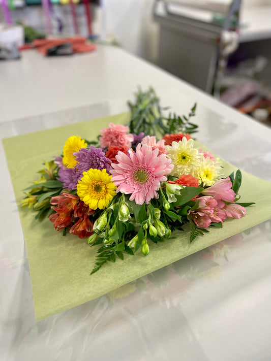 The "Spring Moments" bouquet by Posies Flowers & Fashion features a colorful arrangement of various flowers, including pink gerbera daisies, yellow daisies, and purple chrysanthemums, spread out on green wrapping paper on a white table. Greenery is interspersed among the flowers for added texture and color contrast. Please note that flower availability may vary.