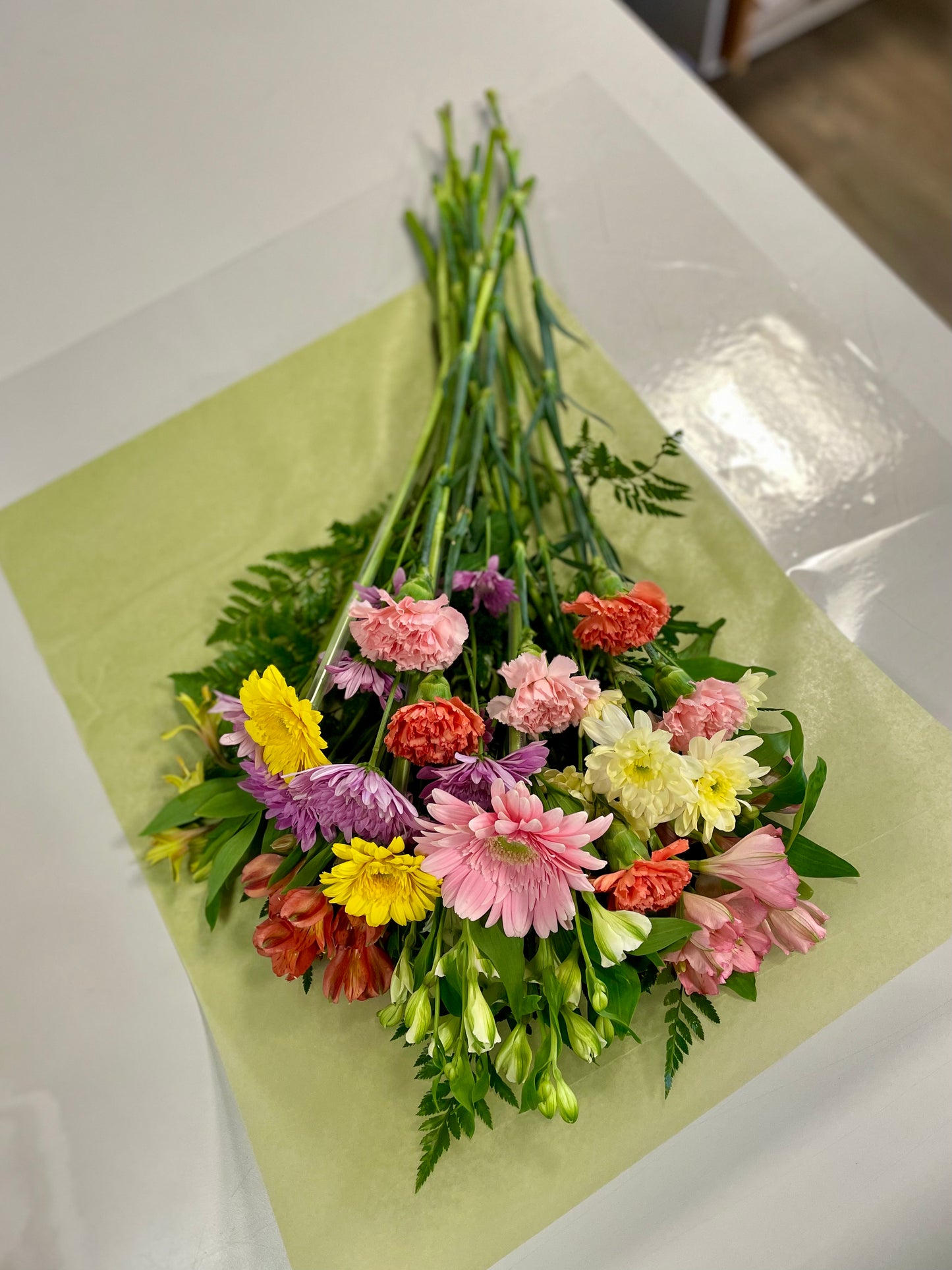 The "Spring Moments" bouquet by Posies Flowers & Fashion features a colorful arrangement of various flowers, including pink gerbera daisies, yellow daisies, and purple chrysanthemums, spread out on green wrapping paper on a white table. Greenery is interspersed among the flowers for added texture and color contrast. Please note that flower availability may vary.