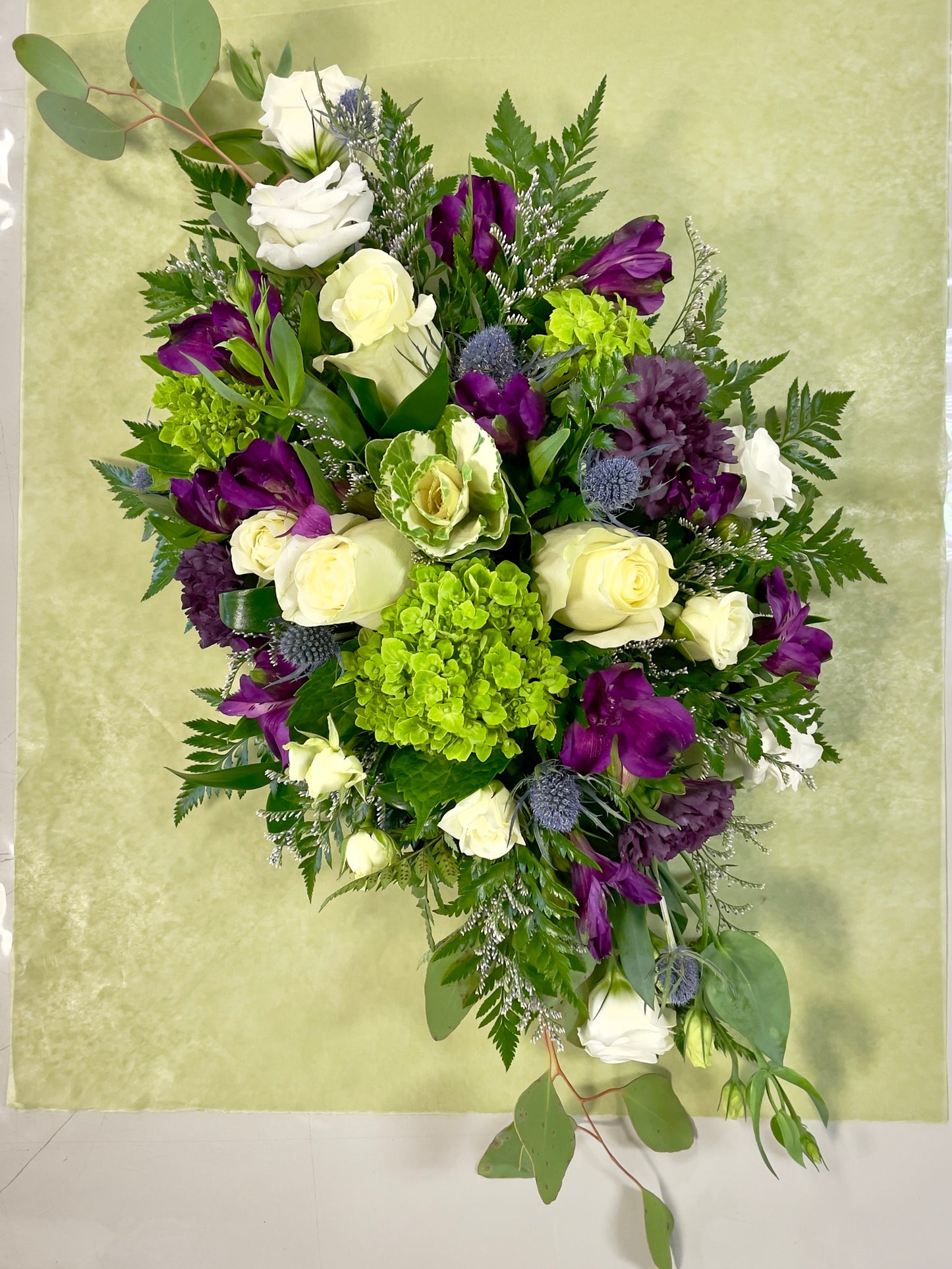 Introducing the "Garden of Royalty Centrepiece" by Posies Flowers & Fashion, showcasing a stunning mix of purple tulips, white roses, and green hydrangeas. Accented with greenery and ferns, this elegant floral arrangement is displayed on a light green placemat atop a white table. This exquisite piece enhances the room's simplicity, against a plain white wall with a partially visible window frame.