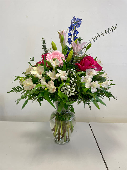 The "Floral Hugs" arrangement by Posies Flowers & Fashion showcases a vibrant mix of pink roses, white lilies, green foliage, and blue delphiniums in a clear glass vase against a plain white background. The flowers and greenery cascade naturally, creating a lush and balanced display; substitutes may be used depending on flower availability.