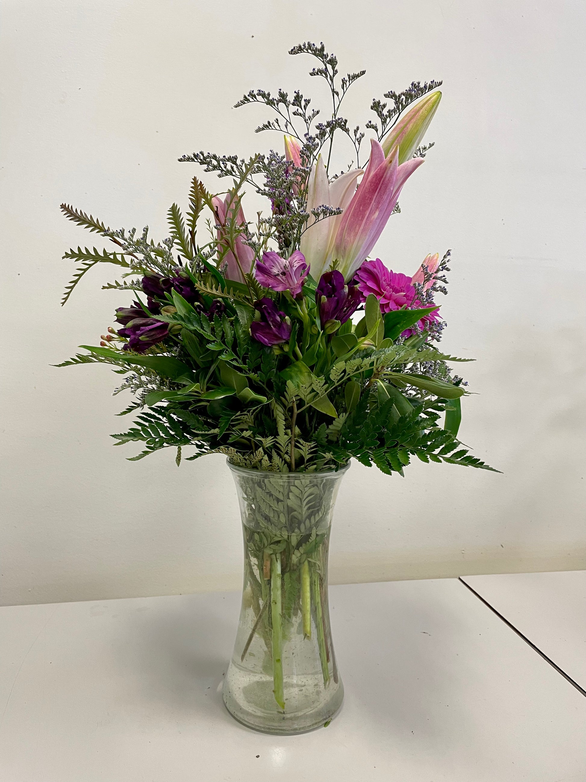 The "Reach for the Sky" bouquet from Posies Flowers & Fashion features a clear glass vase filled with pink lilies, purple daisies, and various types of greenery. Flower substitutions may occur based on availability. The arrangement is set against a plain, light-colored wall backdrop.