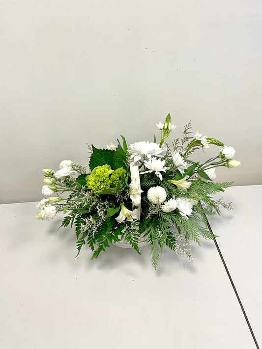 Refined Whites by Posies Flowers & Fashion is an elegant floral arrangement, showcasing white carnations and bellflowers complemented by green hydrangea and assorted greenery, all presented in a sleek white rectangular container. The arrangement is set on a plain white surface against a light-colored wall.