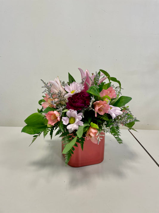 The "Small & Sweet" floral arrangement by Posies Flowers & Fashion features a delightful mix of pink, white, and burgundy flowers accentuated with lush green foliage, beautifully presented in a square red vase. This charming bouquet is showcased on a light-colored surface against a plain background. Please note that due to availability, some flowers may be substituted to maintain the same stunning effect.