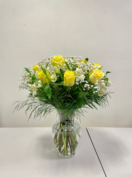 The Kisses of Sunshine Vase by Posies Flowers & Fashion showcases yellow roses, white flowers, and greenery in a clear vase. Substitutions may occur due to availability. The elegant bouquet is beautifully presented against a simple white backdrop on a table for an ideal product image.