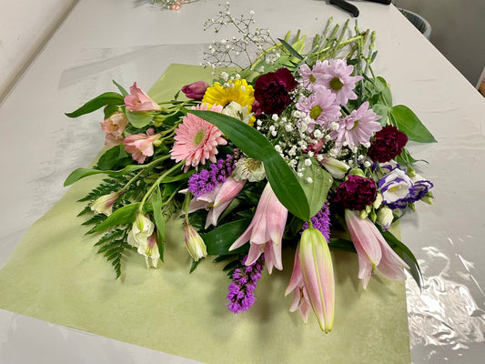 Introducing "Mother’s Delight" by Posies Flowers & Fashion: a vibrant bouquet featuring an exquisite mix of pink lilies, white lilies, pink gerberas, red carnations, yellow flowers, purple flowers, and airy sprigs of baby's breath. The blossoms are beautifully arranged on a light green and white surface and surrounded by lush green leaves. Please note that substitutions may occur based on availability.