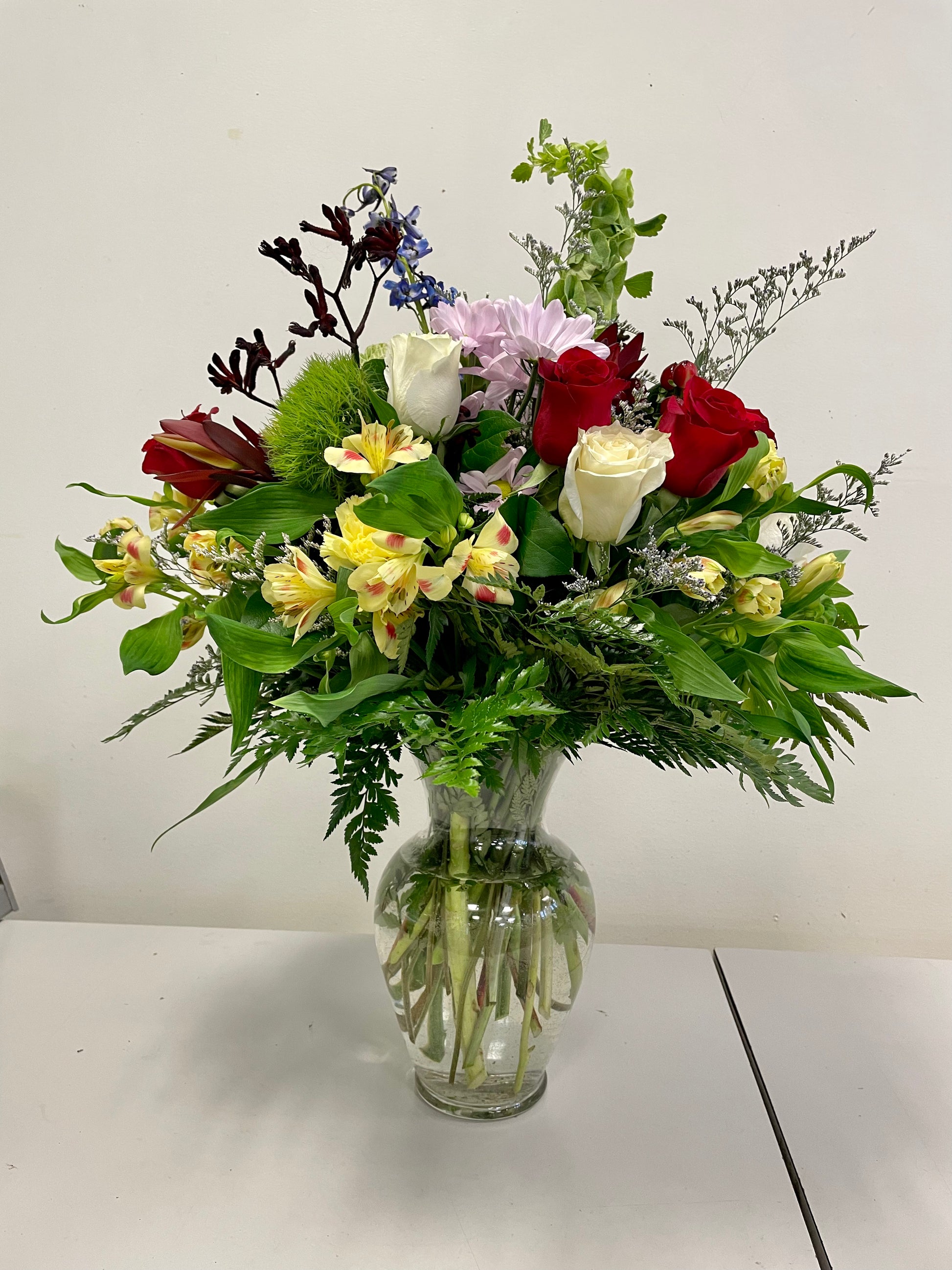 The "Love & Wonder" bouquet from Posies Flowers & Fashion is presented in a clear glass vase, showcasing a vibrant assortment of flowers including red roses, yellow carnations, purple chrysanthemums, and blue delphiniums. Accented with green foliage and ferns for an accurate depiction, this stunning arrangement is displayed on a white table against a plain white wall.