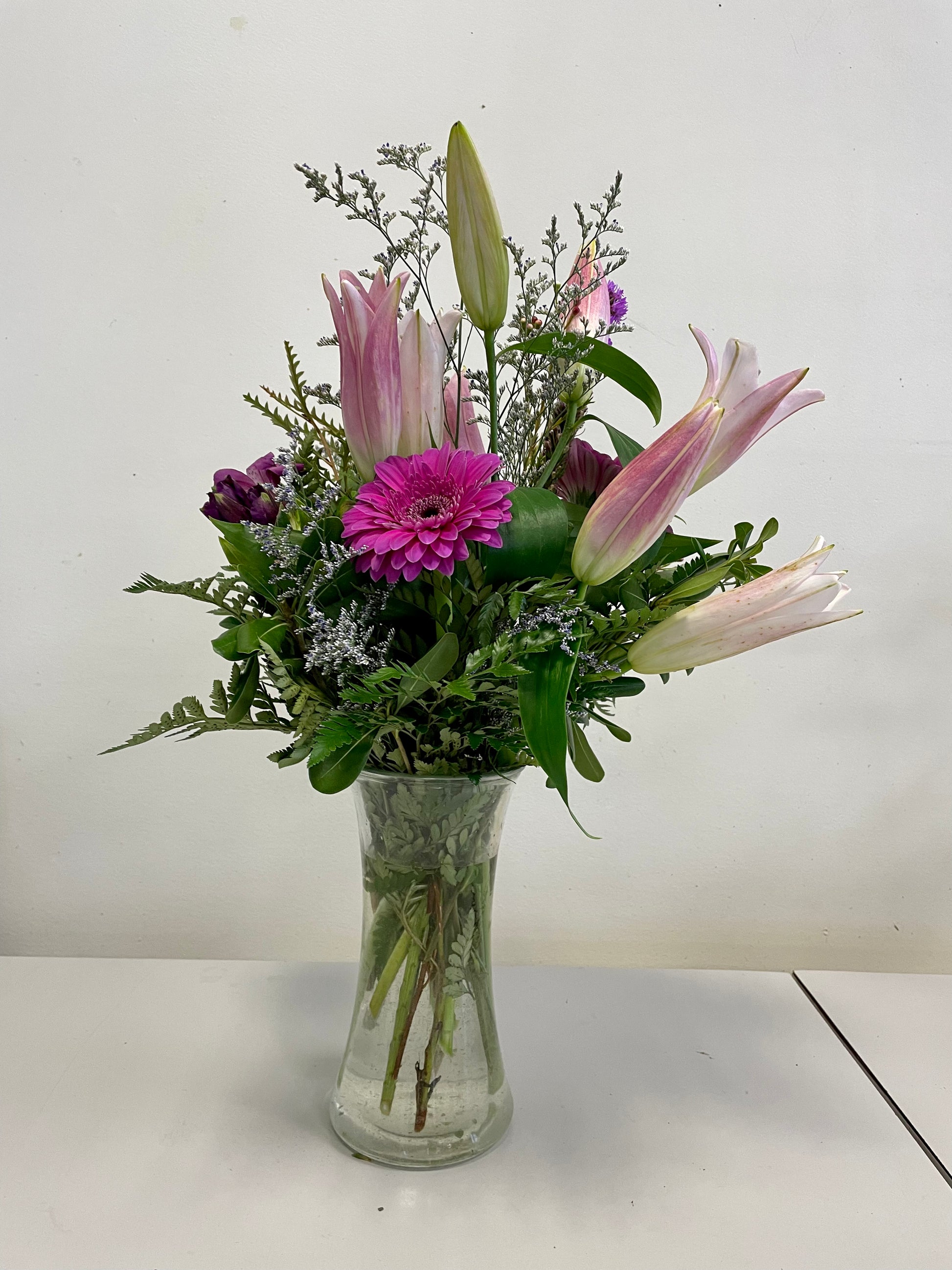 The "Reach for the Sky" bouquet from Posies Flowers & Fashion features a clear glass vase filled with pink lilies, purple daisies, and various types of greenery. Flower substitutions may occur based on availability. The arrangement is set against a plain, light-colored wall backdrop.