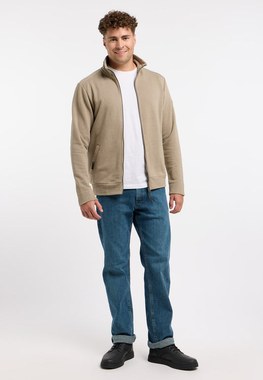 A person with curly hair is smiling and standing against a plain white background. They are wearing the Miet Sweatshirt by RAGWEAR, a comfortable beige sweatshirt, over a white shirt, paired with versatile blue jeans and black shoes.