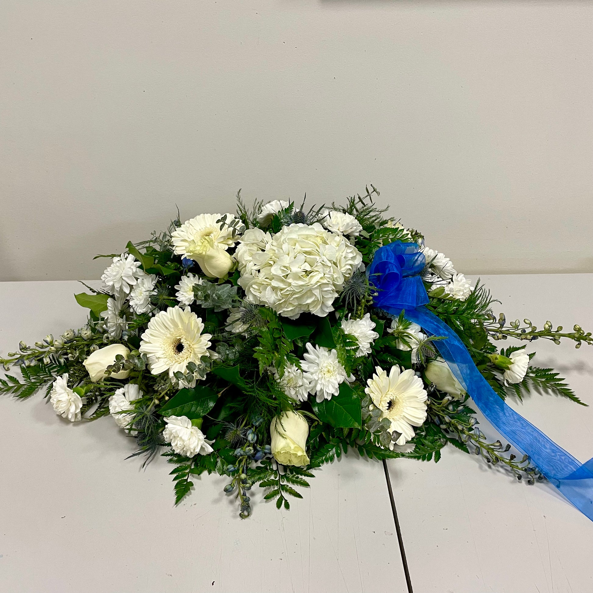 The Simple Whites arrangement from Posies Flowers & Fashion features a beautiful mix of white roses, chrysanthemums, and daisies, accented with lush green leaves and fern fronds. Displayed elegantly on a white surface against a plain light-colored wall, this floral design is interspersed with delicate white hydrangeas and adorned with a blue ribbon tied on the right side.