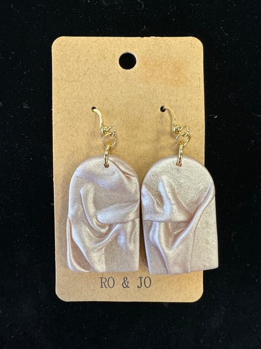 Introducing the Ro&Jo Earrings 428 by RO&JO: a stunning pair of pearl-colored, handmade polymer clay earrings featuring an abstract, folded design that elegantly mimics draped fabric. These exquisite earrings are attached to a beige cardboard backing, complete with a convenient hanging hole at the top and "RO & JO" printed at the bottom. Set against a black background, these pieces truly shine as remarkable examples of artisan jewelry.