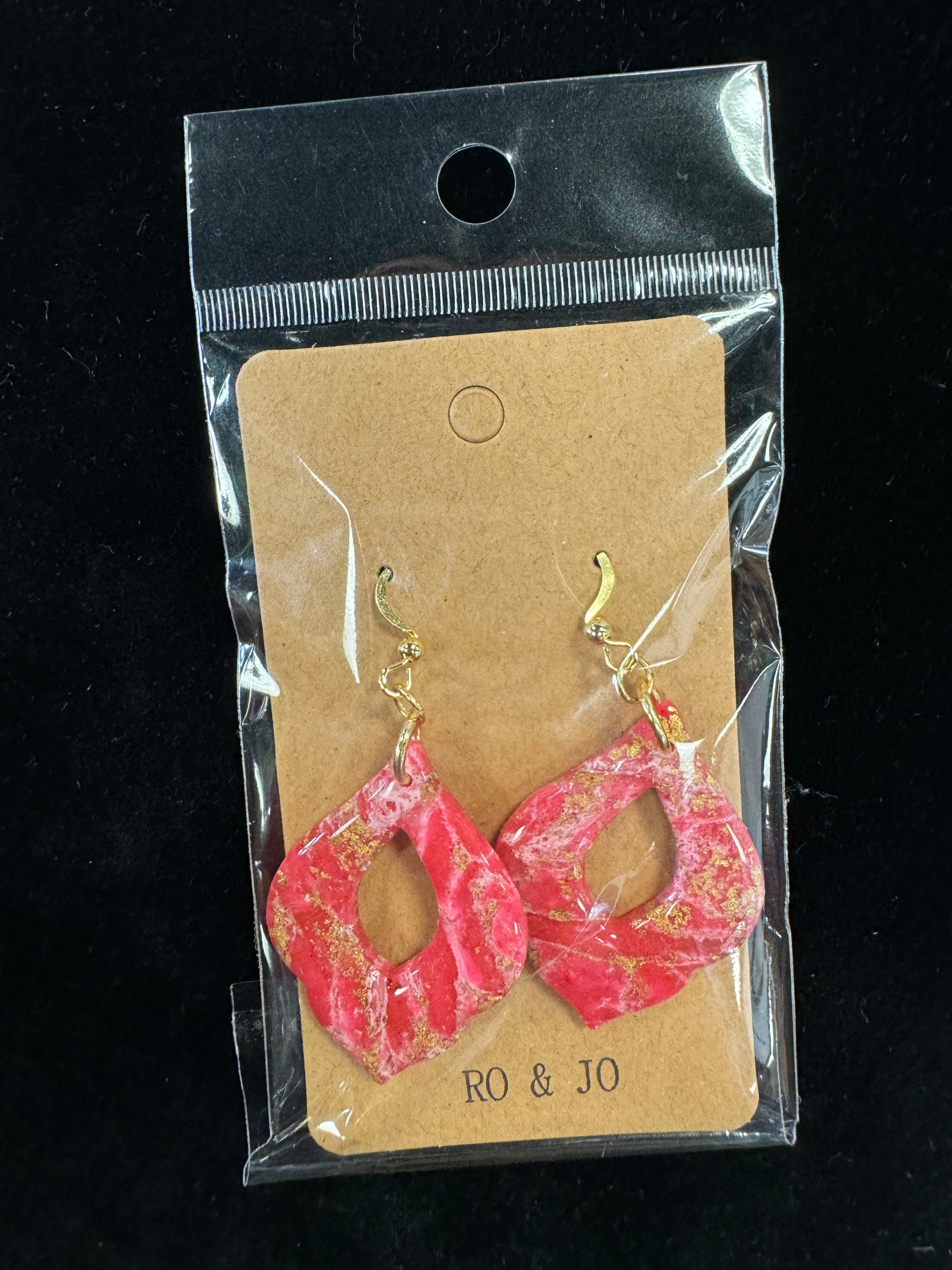 A pair of vibrant red and pink marbled polymer clay earrings with gold accents, showcasing an abstract, cut-out geometric design. The handmade Ro&Jo Earrings 446 from RO&JO are displayed on a kraft paper backing labeled "RO & JO" and encased in clear plastic packaging against a black background.