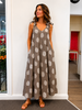 A woman stands indoors wearing the ETERNELLE Linen Jumpsuit Taupe 6-SS26168-9, a loose brown sleeveless jumpsuit with large white polka dots and tied shoulder straps, smiling with her hands in her pockets.