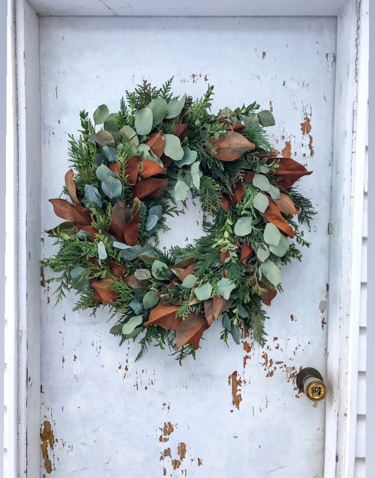 A Mixed Greens with Eucalyptus Wreath from Posies Flowers & Fashion, featuring a blend of natural greens and earthy reddish-brown leaves, adorns a weathered white door with chipped paint. The subtle gleam of the brass doorknob introduces an elegant touch to this charming display against a light-colored wall.