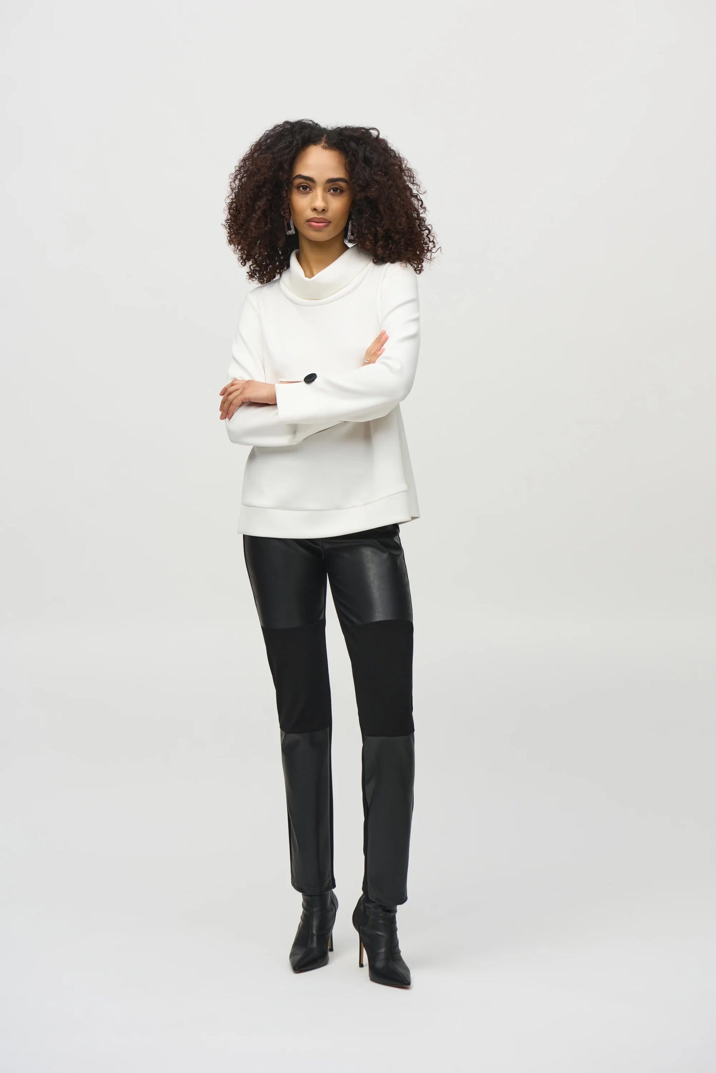 A woman with curly hair wears a loose-fitting, long-sleeved white Scuba Jersey Boxy Top 244129 by Joseph Ribkoff. The top, featuring button details on the sleeves, lends a boxy silhouette. She stands in front of a plain background, looking slightly downward with a neutral expression.