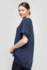 A person with long hair tied back stands sideways, smiling over their shoulder. They're wearing the loose-fitting TOP CS25-609 UCHUU Montreal dark blue blouse with satin trim and black pants. The background is a plain light gray.