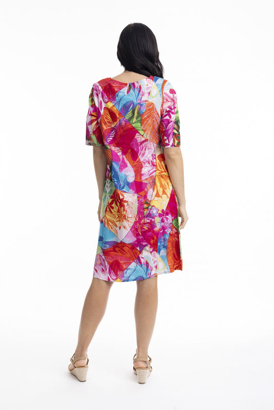 A woman with long dark hair wears the ORIENTIQUE Jozani Contemporary Dress Style 30057, featuring a vibrant multicolored tropical leaf print. She pairs it with light heeled sandals against a plain white background.