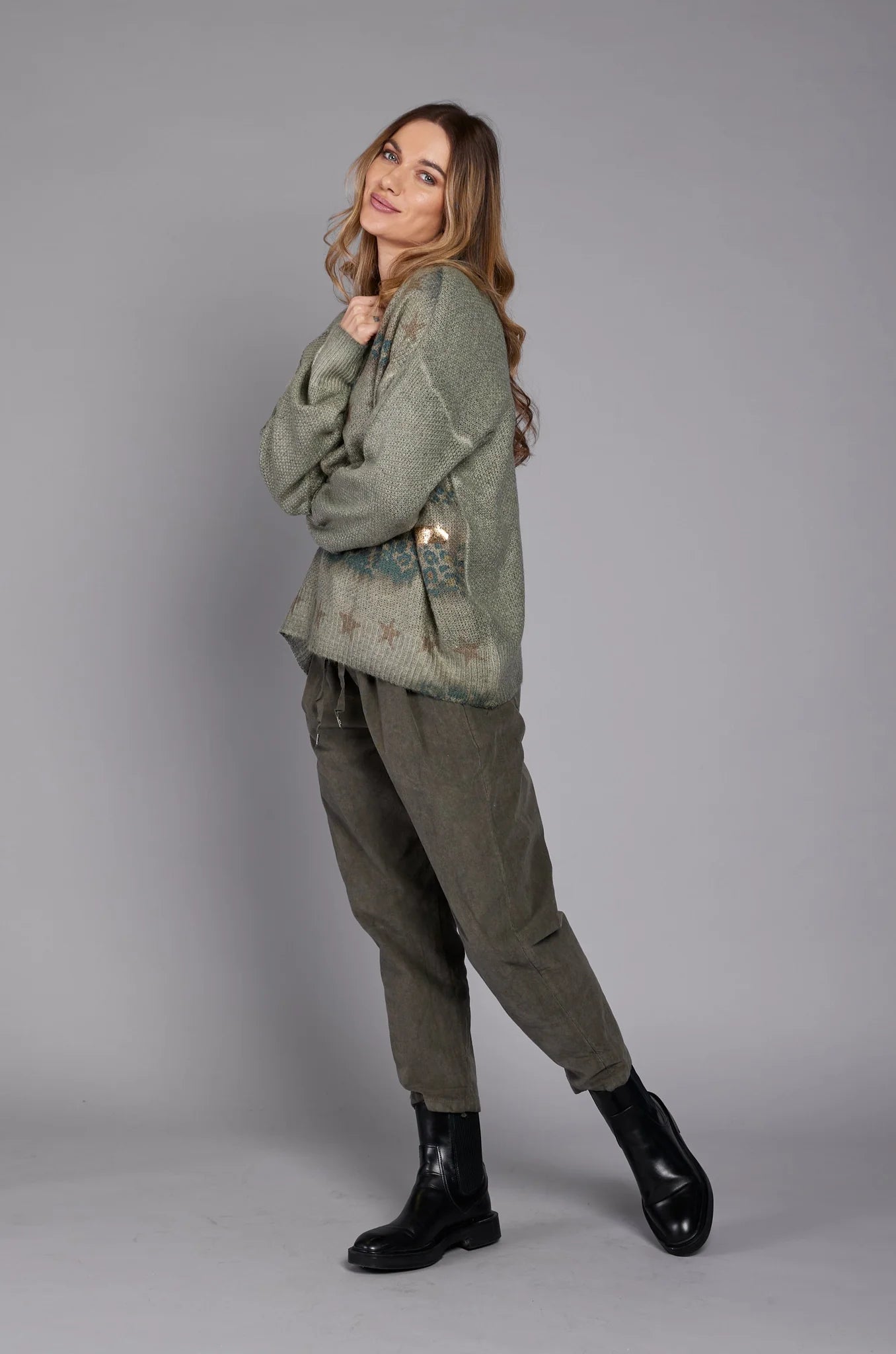 A person wearing a gray long-sleeve top, the stylish EL27901 pants by ELISSIA in ankle-high olive green with a drawstring, and black ankle boots stands with their hands in their pockets against a plain grey background. A scarf is draped over their shoulder. Only the lower half of the person is visible.