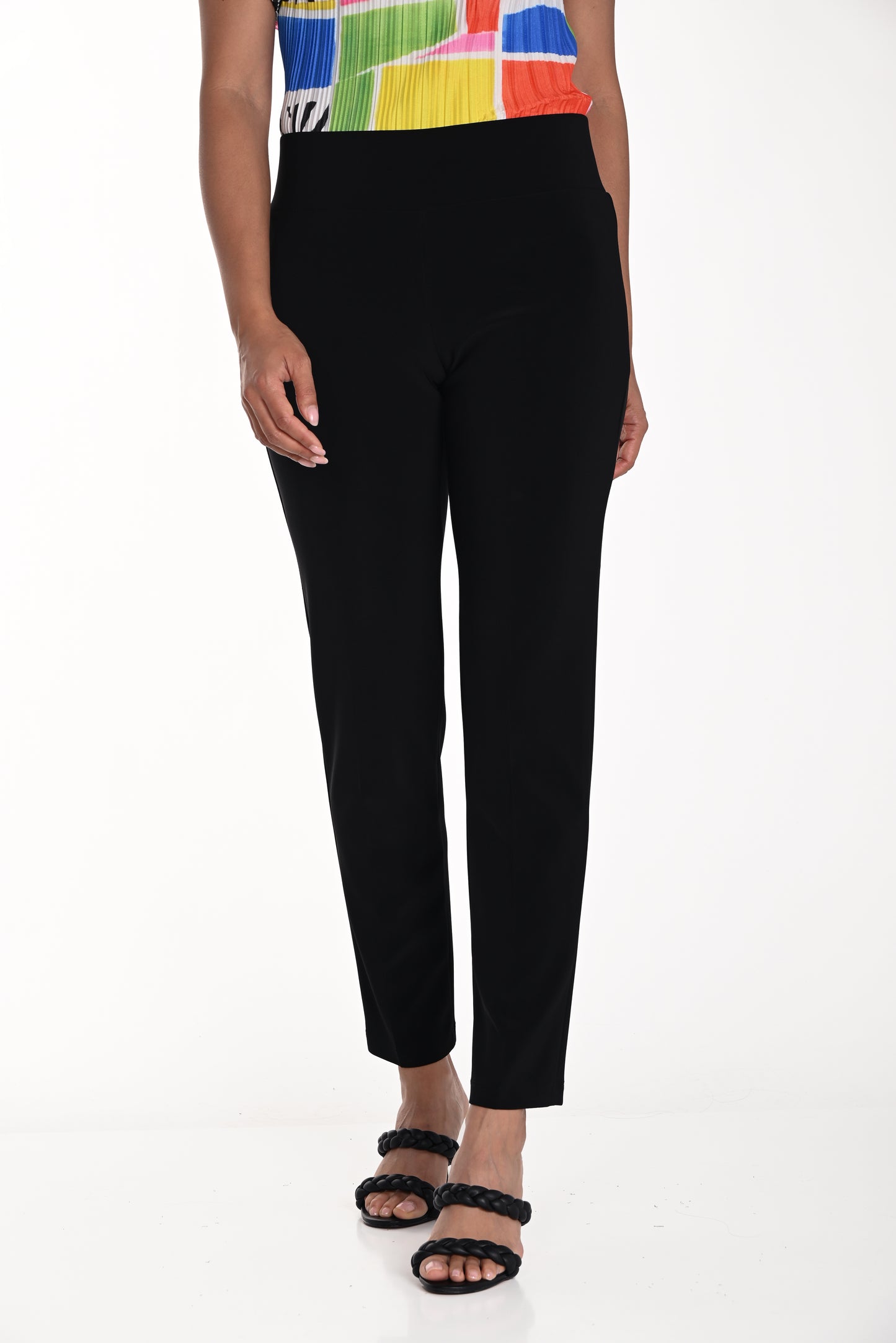 A person, visible from the waist down against a white background, wears the Frank Lyman Slim Leg Pull-On Pant in Black (Style 082)—a stylish Canadian-made pant—paired with a colorful sleeveless top and black braided sandals.