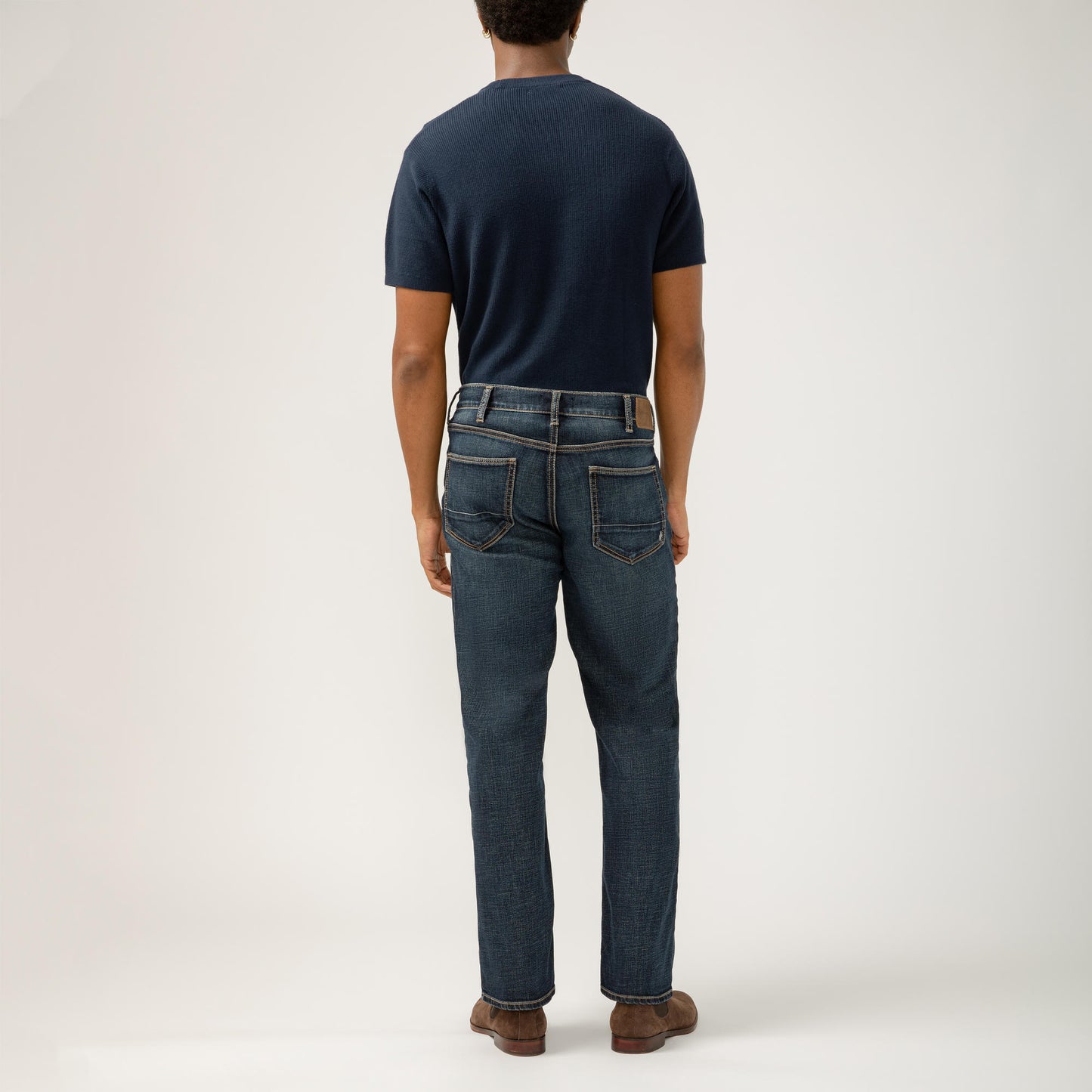 A person stands against a plain background wearing a navy blue, classic fit, short-sleeve shirt tucked into Grayson Classic Fit Straight Leg Silver Jeans M23425EFS409 by Silver Jeans Co. with brown suede shoes. Their arms rest comfortably by their sides, and their shoes contrast with the white backdrop. Style code: SIL-M23425EFS409-IND.