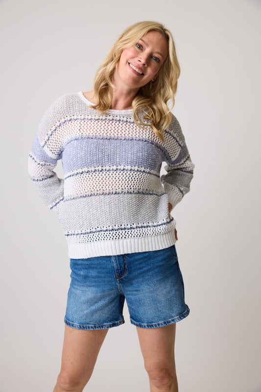 A person with long blonde hair smiles in a Harper Eco Stripe 75337 sweater by Parkhurst, crafted from sustainable materials. The light-colored knit features blue and gray stripes, paired with denim shorts against a plain light background, creating a versatile wardrobe look.