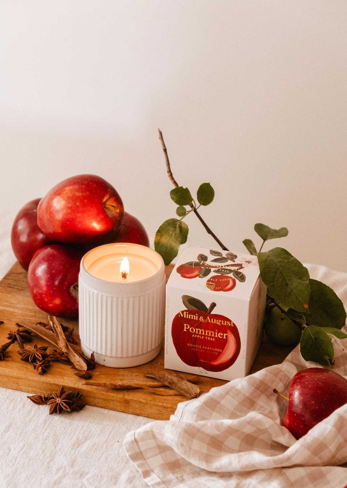 A glowing Pommier – Reusable Soy Candle (8 oz) by Mimi & August rests on a wooden board, surrounded by red apples, star anise, and its branded box. Green leaves and a beige cloth add to the warm, autumn atmosphere.