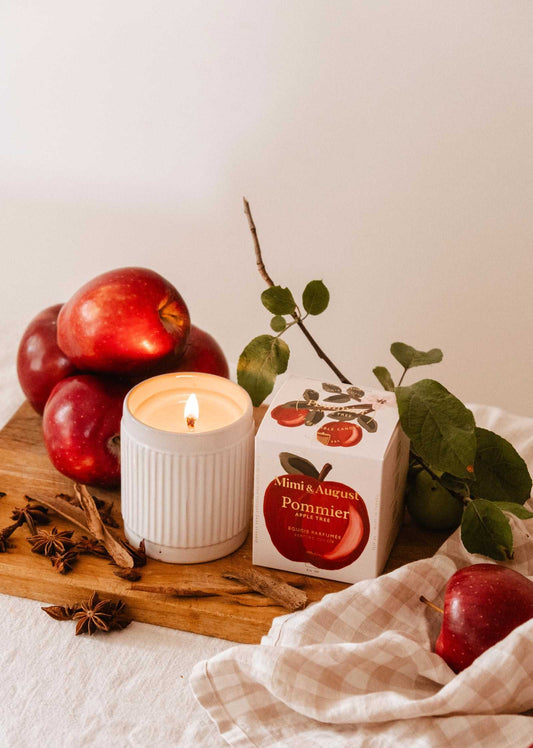 A glowing Pommier – Reusable Soy Candle (8 oz) by Mimi & August rests on a wooden board, surrounded by red apples, star anise, and its branded box. Green leaves and a beige cloth add to the warm, autumn atmosphere.