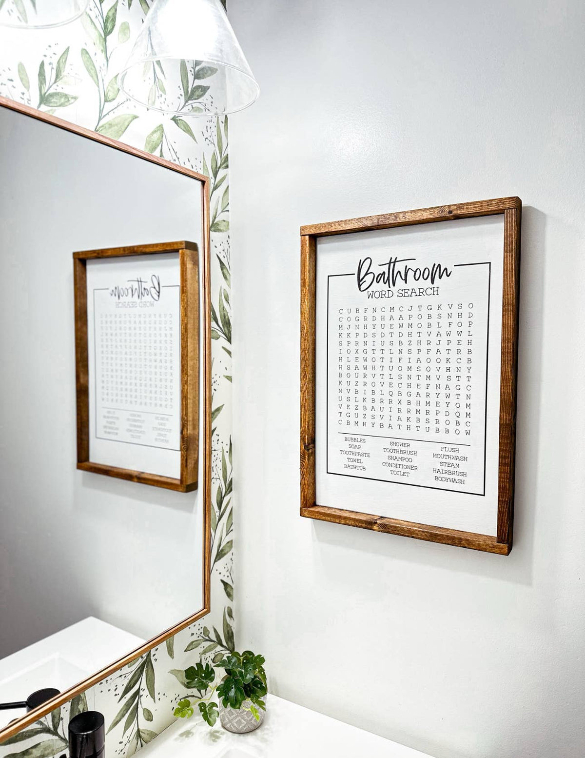 An Inspired Findings “Bathroom Wordsearch” wooden framed sign hangs on a white wall above the sink, with part of the mirror and some greenery visible in this stylish, modern bathroom.