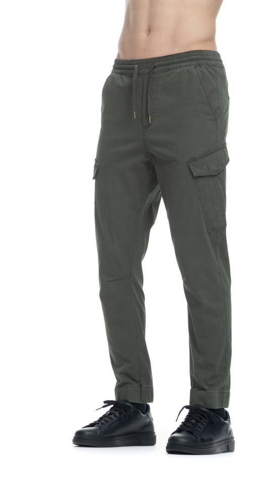 A cropped image shows a person wearing Zevl Dark Oliven Trousers by RAGWEAR, featuring a drawstring waist and side pockets, paired with black sneakers. The person's torso is not visible, and the vegan trousers have a casual, relaxed fit perfect for any casual occasion.