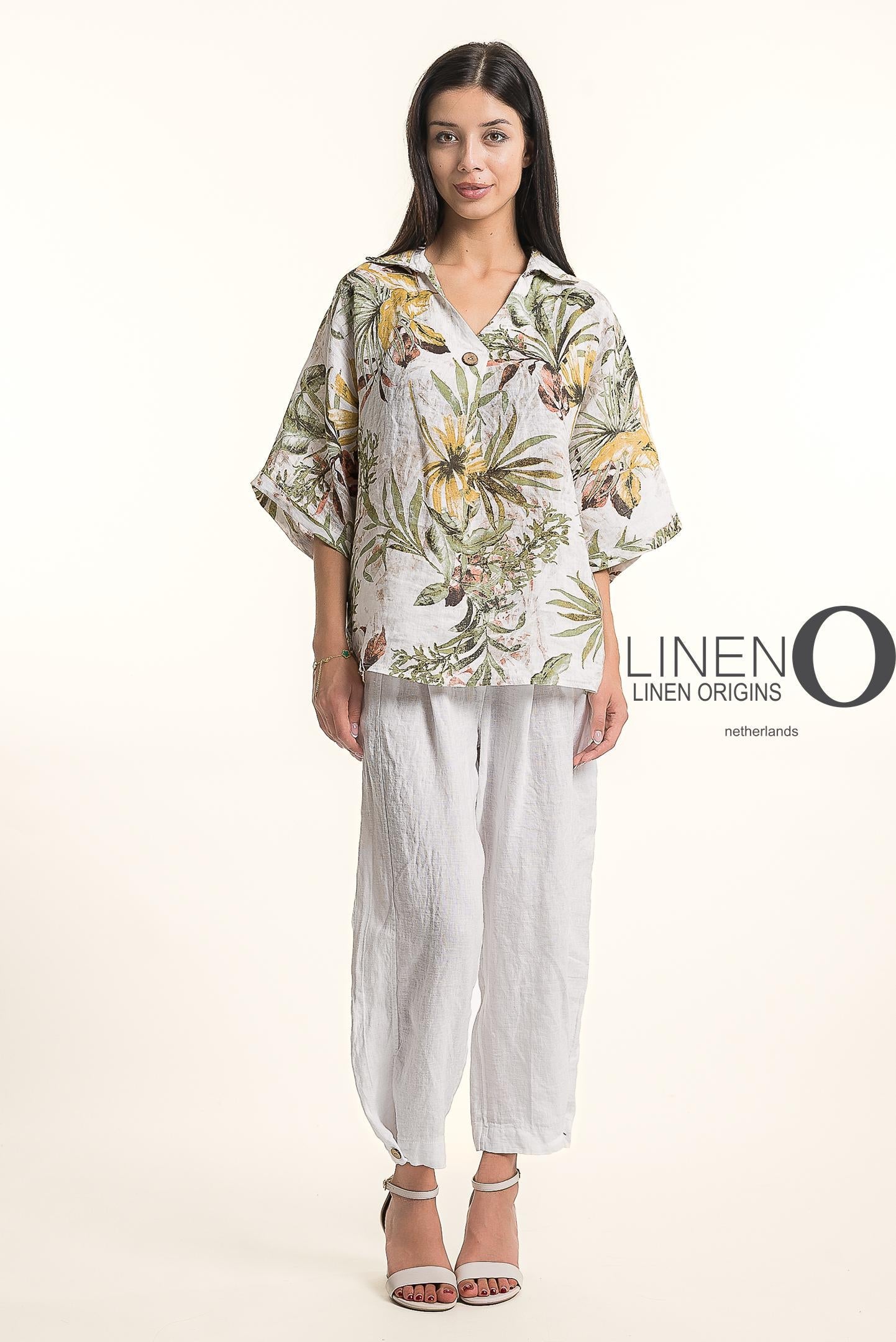 A woman wears the LinenO French Linen Blouse TP1714P-11 Bianco, featuring a loose fit and floral pattern, paired with white linen pants and open-toed sandals. She has long dark hair. The background is plain white with "LINEN ORIGINS.